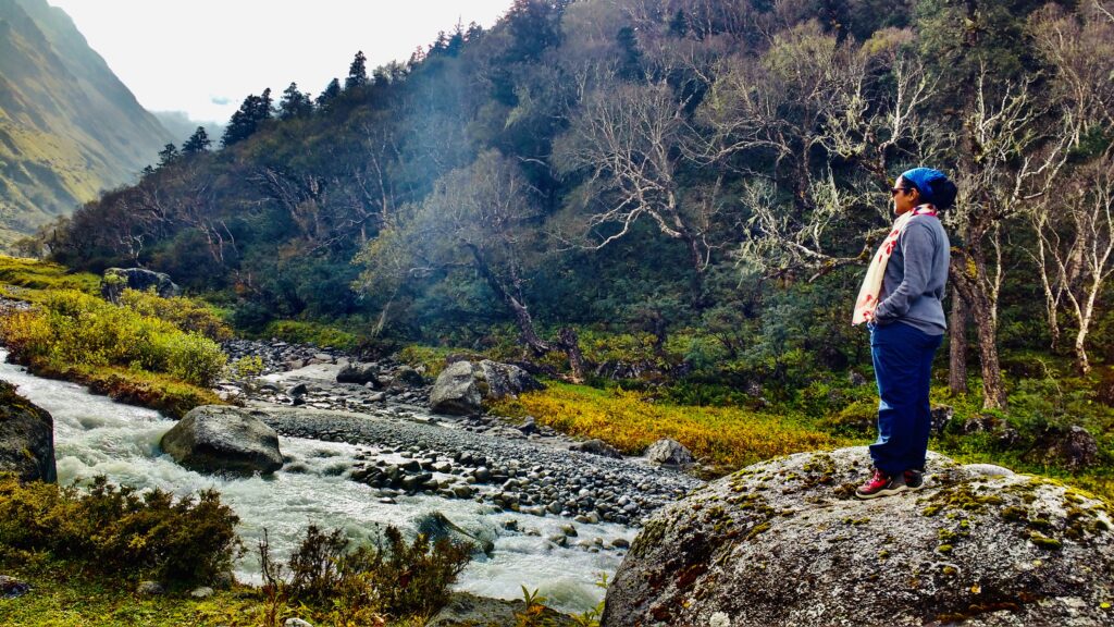 A girl standing on a rock looking at a beautiful view of mountains and river at the summit of Har ki doon trek in Uttarakhand, India